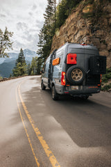 Gray off-road camper van on a scenic mountain road with rocky cliffs and snow-capped mountains. Perfect for your RV rental adventure.