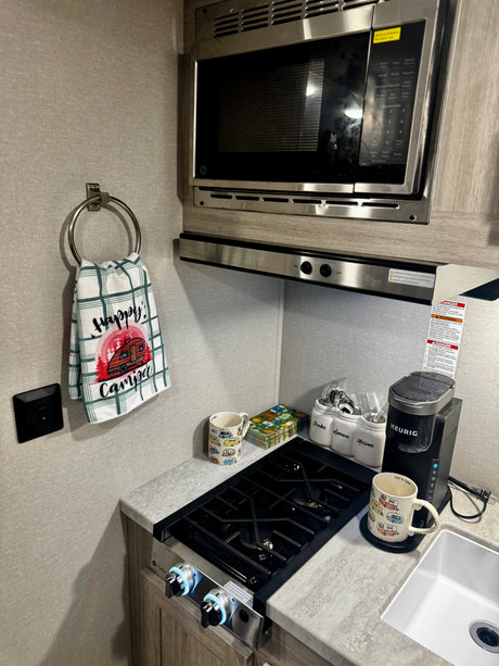 This RV rental kitchen features a microwave, stovetop, Keurig coffee maker, and "Happy Camper" decor, offering compact convenience.
