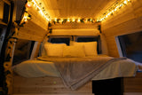 Cozy RV rental bedroom featuring wood walls, string lights with greenery, a sunflower accent, and a plush bed.