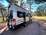 Silver Coachmen camper van (RV rental) with open door & bike rack, parked on gravel amidst sunny pine trees. Ready for nature.
