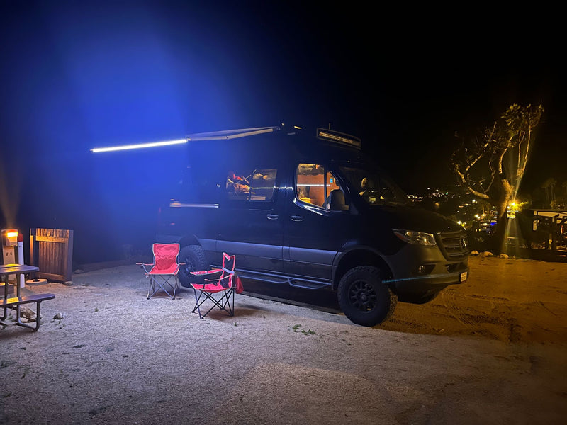 RV rental adventure: A dark camper van lights up the night with its awning, red chairs, and picnic table on gravel. — photo 16