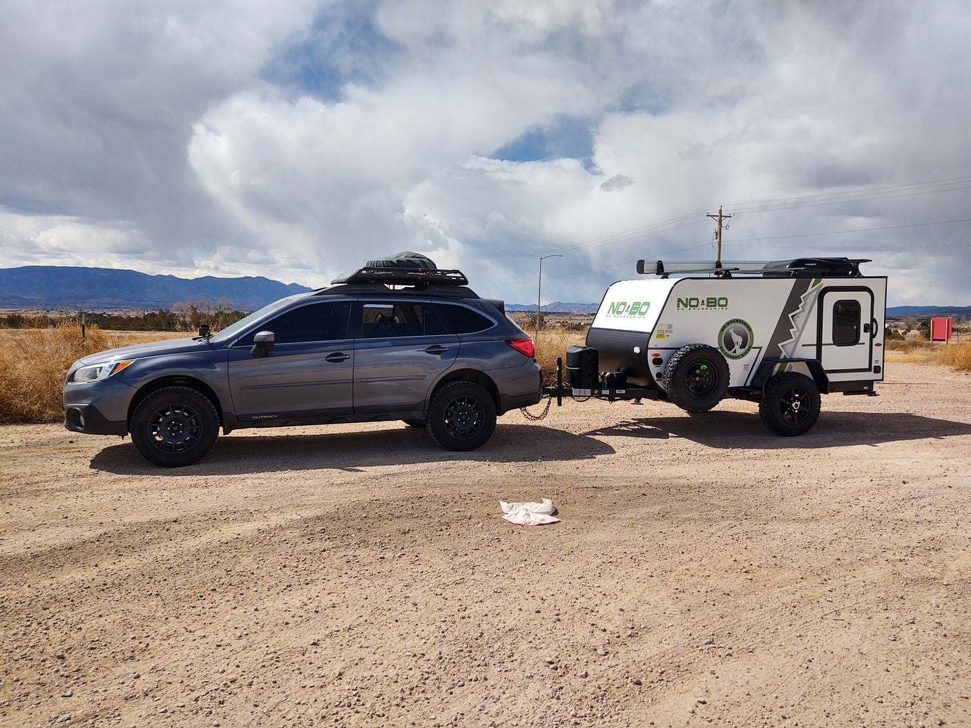 A Subaru Outback tows a compact NO BO camper on a dirt road.