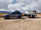 A Subaru Outback tows a compact NO BO camper on a dirt road.