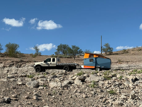 RV rental adventure! A sturdy truck tows a blue & orange pop-up camper on a rugged, rocky desert trail under blue skies.