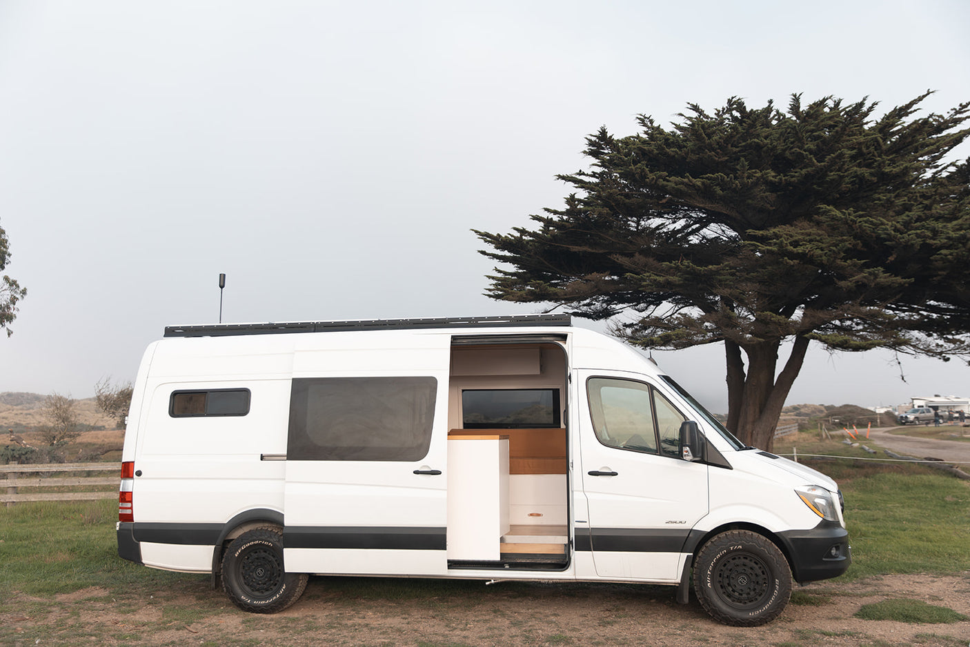 White Sprinter camper van for rv rental, ready for travel, open door reveals custom interior by a tree.