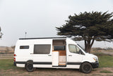 White Sprinter camper van for rv rental, ready for travel, open door reveals custom interior by a tree.