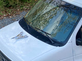 Front of a white van, possibly an RV rental, with a chrome winged hood ornament & wipers. Trees reflect in the windshield.