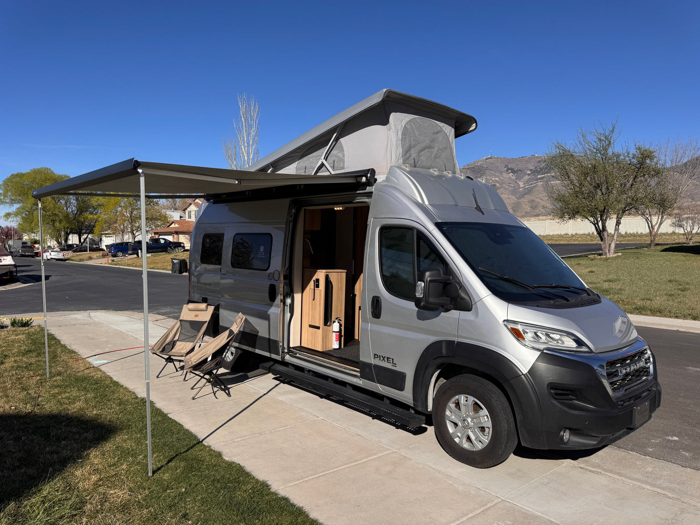 Silver pop-top camper van (rv rental) with awning extended, chairs set out. Interior viewable, parked outdoors with mountains.