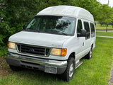 White high-top Ford E-Series conversion van (Transmaster), ideal RV rental, parked on grass by trees on a cloudy day.