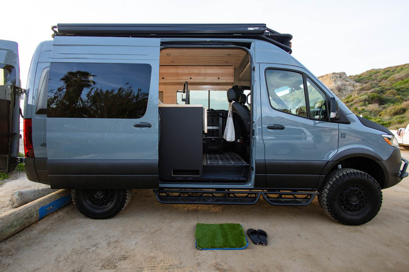 This grey RV rental camper van, ideal for van life, has its side door open showing a modern interior. Flip-flops rest on a mat outside.