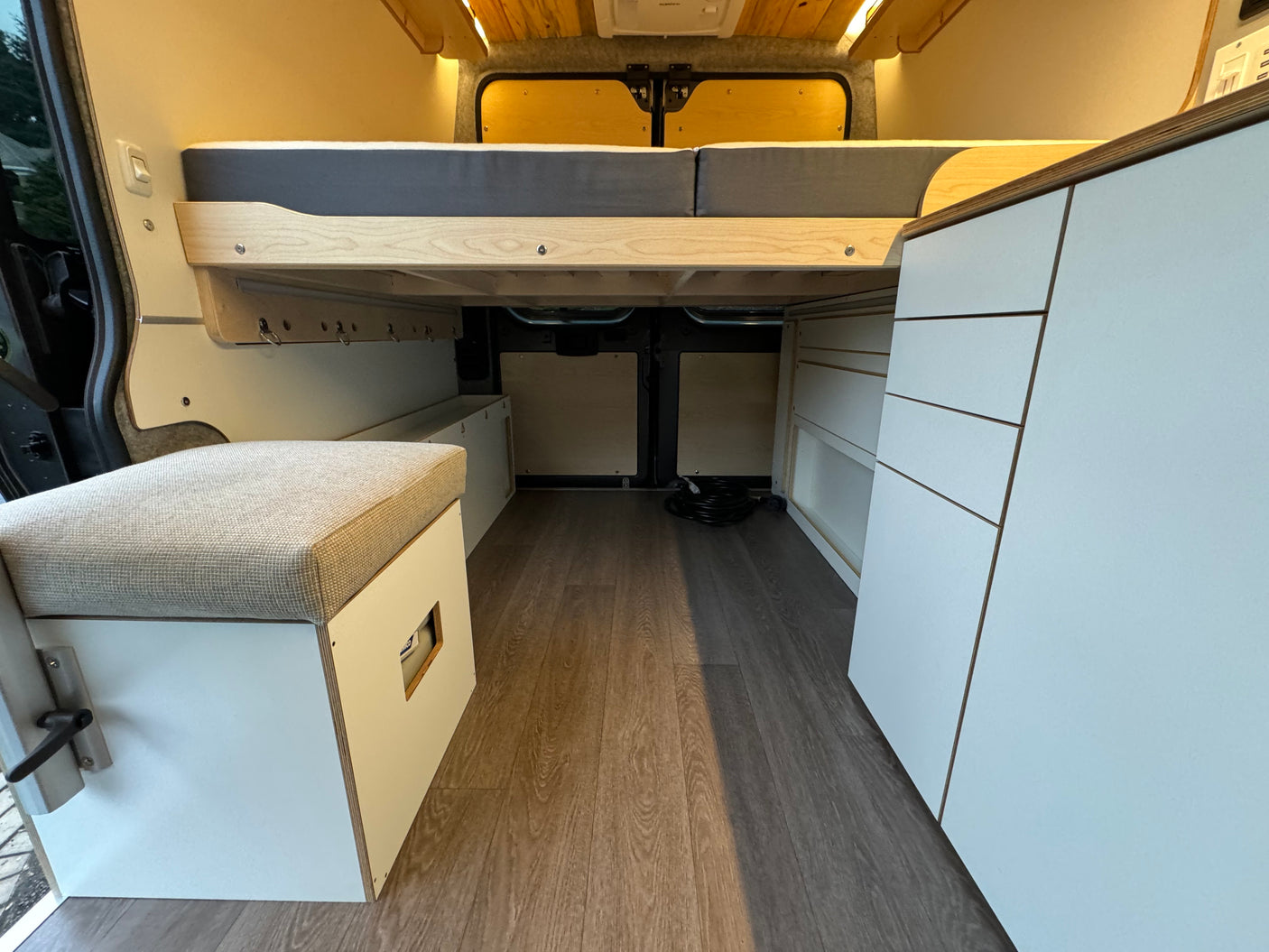Spacious RV rental interior with a cozy bed, bench seating, white cabinetry, and wood flooring.