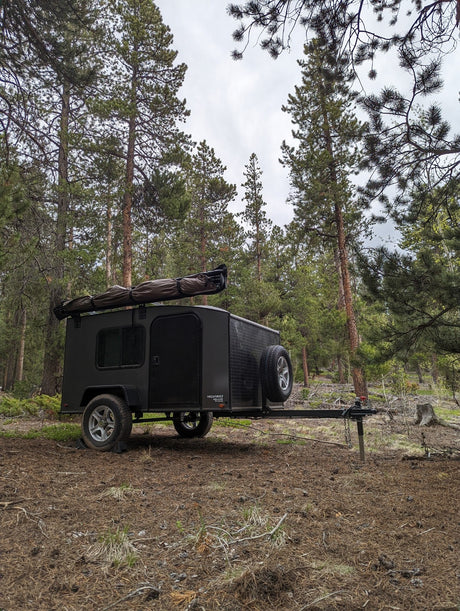 A dark gray off-road teardrop camper, an ideal rv rental, nestled in a serene pine forest for camping.