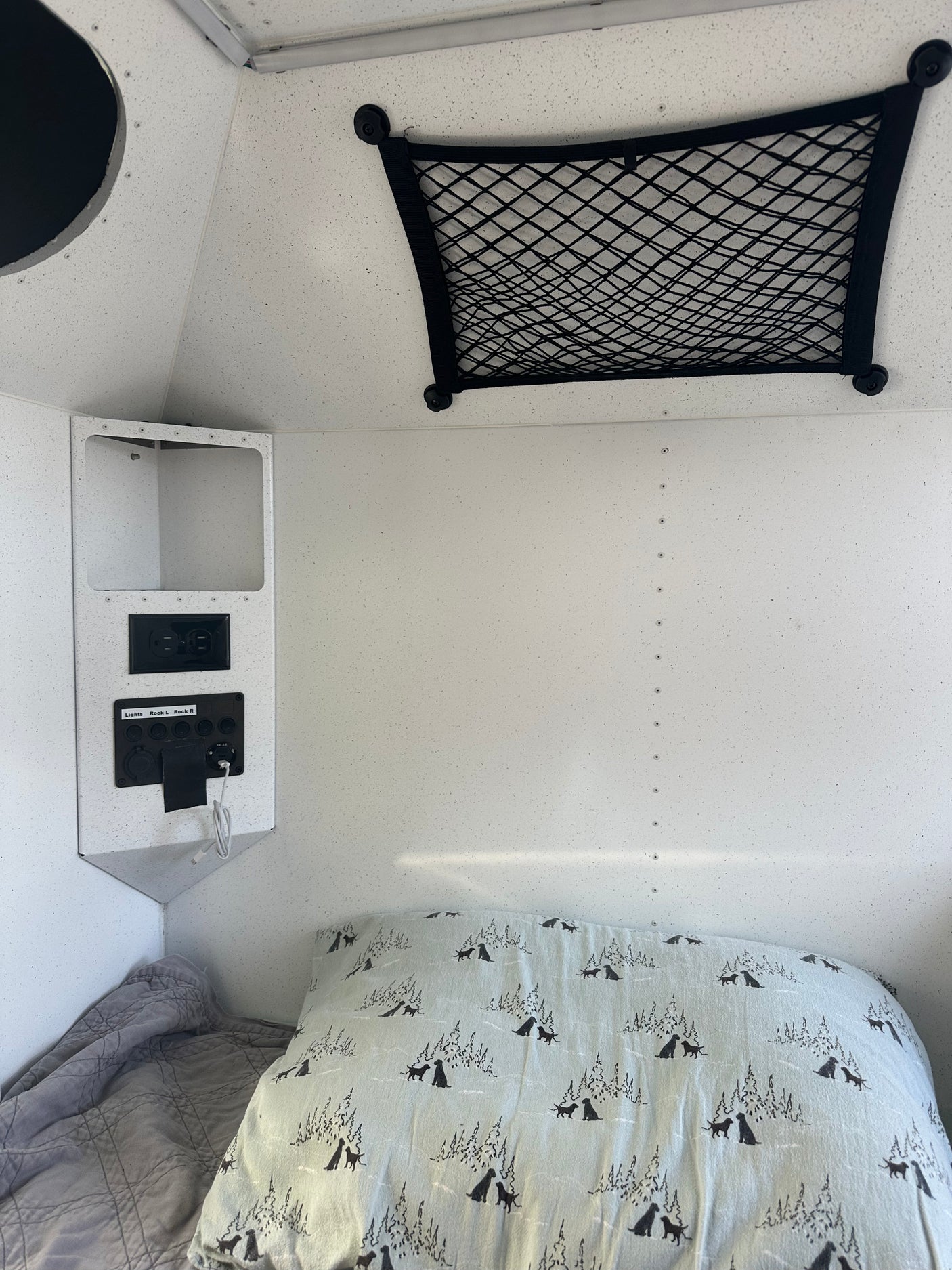 RV rental bed with dog-patterned pillow, power panel (AC/USB outlets), and overhead storage net. Camper interior.