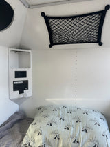 RV rental bed with dog-patterned pillow, power panel (AC/USB outlets), and overhead storage net. Camper interior.