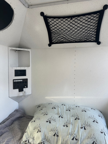 RV rental bed with dog-patterned pillow, power panel (AC/USB outlets), and overhead storage net. Camper interior.