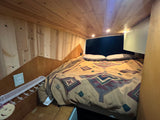 Cozy rv rental bed with patterned blanket, wooden walls/ceiling, recessed lights, and outlet. Ideal camper van sleeping area.