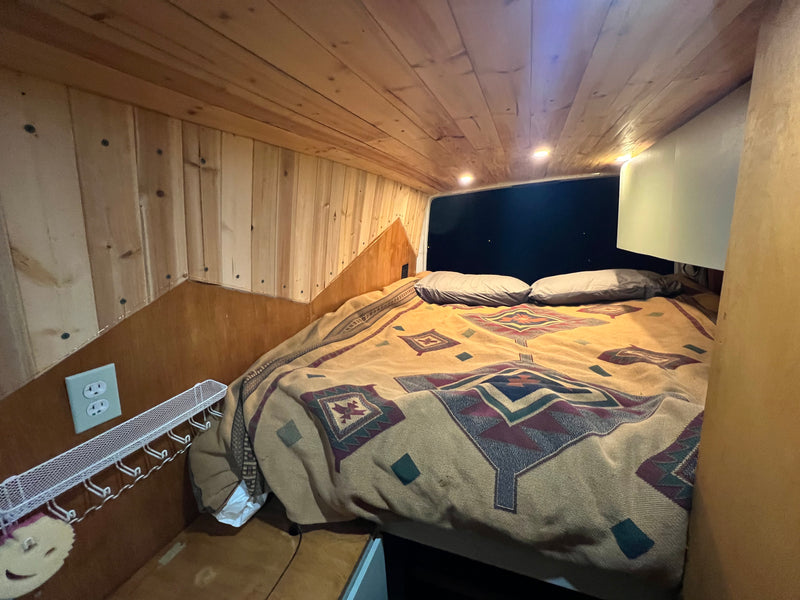 Cozy rv rental bed with patterned blanket, wooden walls/ceiling, recessed lights, and outlet. Ideal camper van sleeping area. — photo 3