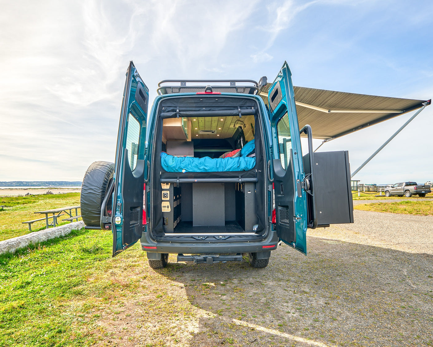 Adventure-ready teal RV rental. Open back reveals a cozy bed, organized storage, and extended awning for beachside relaxation. — photo 4
