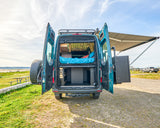 Adventure-ready teal RV rental. Open back reveals a cozy bed, organized storage, and extended awning for beachside relaxation.