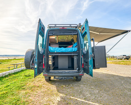 Adventure-ready teal RV rental. Open back reveals a cozy bed, organized storage, and extended awning for beachside relaxation.