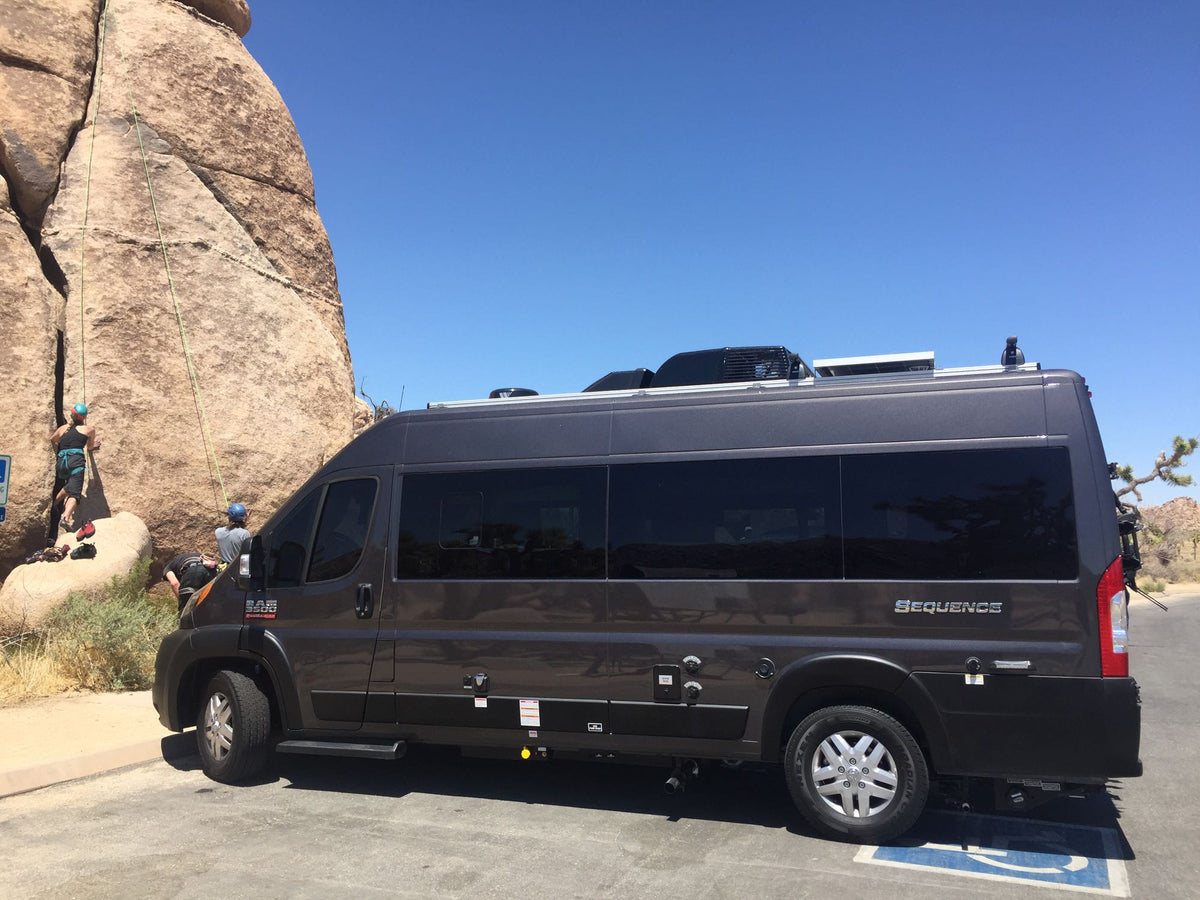 Grey Sequence camper van RV rental by rock climbers in a sunny desert. Your next adventure awaits!|A dark grey Ram ProMaster RV rental is parked on an asphalt path in a sunny park