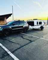 Dark SUV with roof racks hitches to a white teardrop RV rental, a Luna Rover, in a parking lot at sunset.