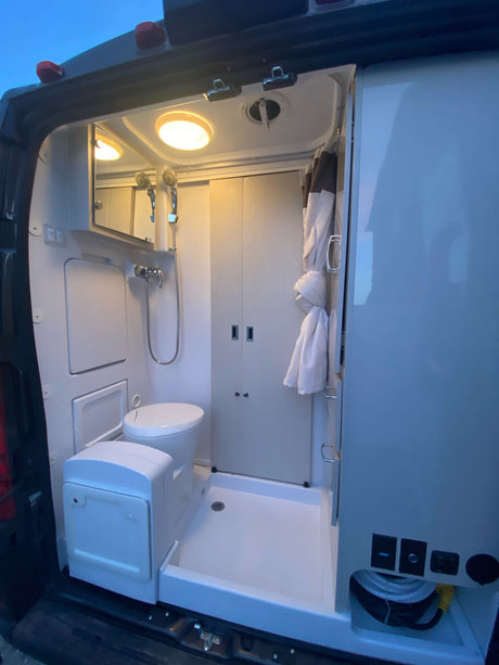 Inside an RV rental, a compact camper van bathroom with a toilet, shower, mirror, and storage, illuminated by overhead lights.