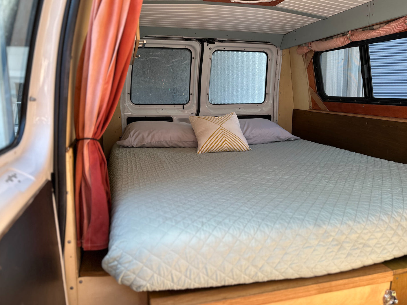 Cozy bed setup inside an RV rental. Features light blue quilted bedding, two pillows, and window privacy coverings in a camper van.
