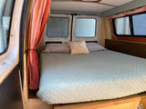 Cozy bed setup inside an RV rental. Features light blue quilted bedding, two pillows, and window privacy coverings in a camper van.