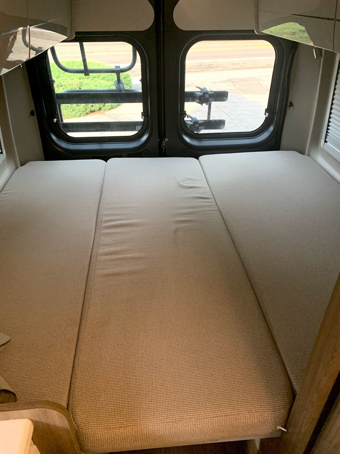 Cozy RV rental camper van interior. A three-panel bed is visible, with rear windows showing a road and bike rack.