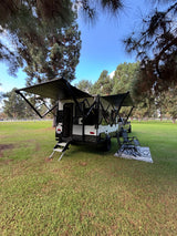 RV rental set up for comfort: a travel trailer with extended awnings, steps, chairs, and a rug on green grass.