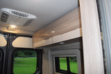 RV rental interior featuring an overhead AC unit, light wood-grain cabinets, and large windows overlooking a green campsite.