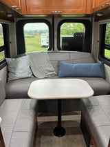 Inside an RV rental: a gray leatherette dinette with a table, decorative pillows, wooden cabinets, and panoramic window views.