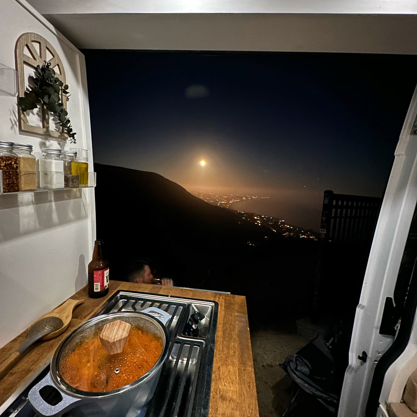 RV rental night: Cozy van kitchen cooking with a bright full moon and city lights view, perfect for van life adventures.