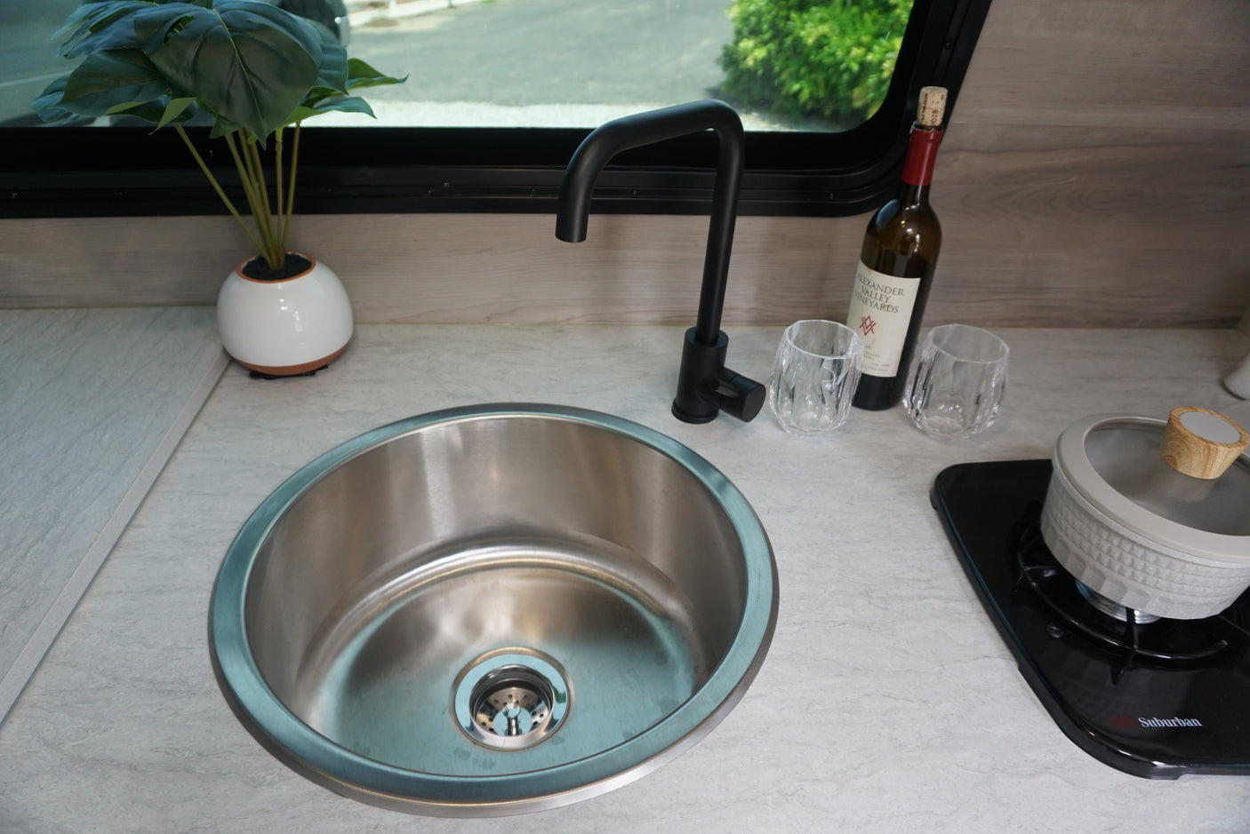 RV rental kitchen: stainless steel sink, black faucet, plant, wine bottle, glasses, and compact stovetop for travel.