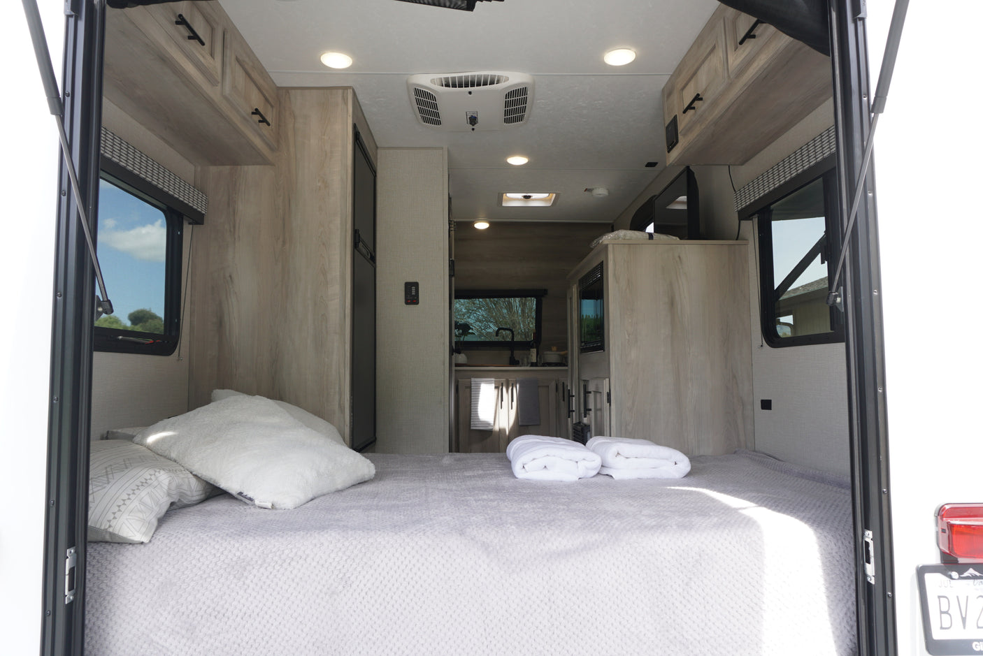 Inside an RV rental: A large, comfortable bed with light grey bedding dominates the foreground, leading to a modern kitchen.