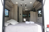 Inside an RV rental: A large, comfortable bed with light grey bedding dominates the foreground, leading to a modern kitchen.
