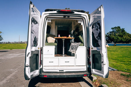 RV rental camper van's open rear doors display its cozy interior, including a table, seating, and world maps, by a scenic park.