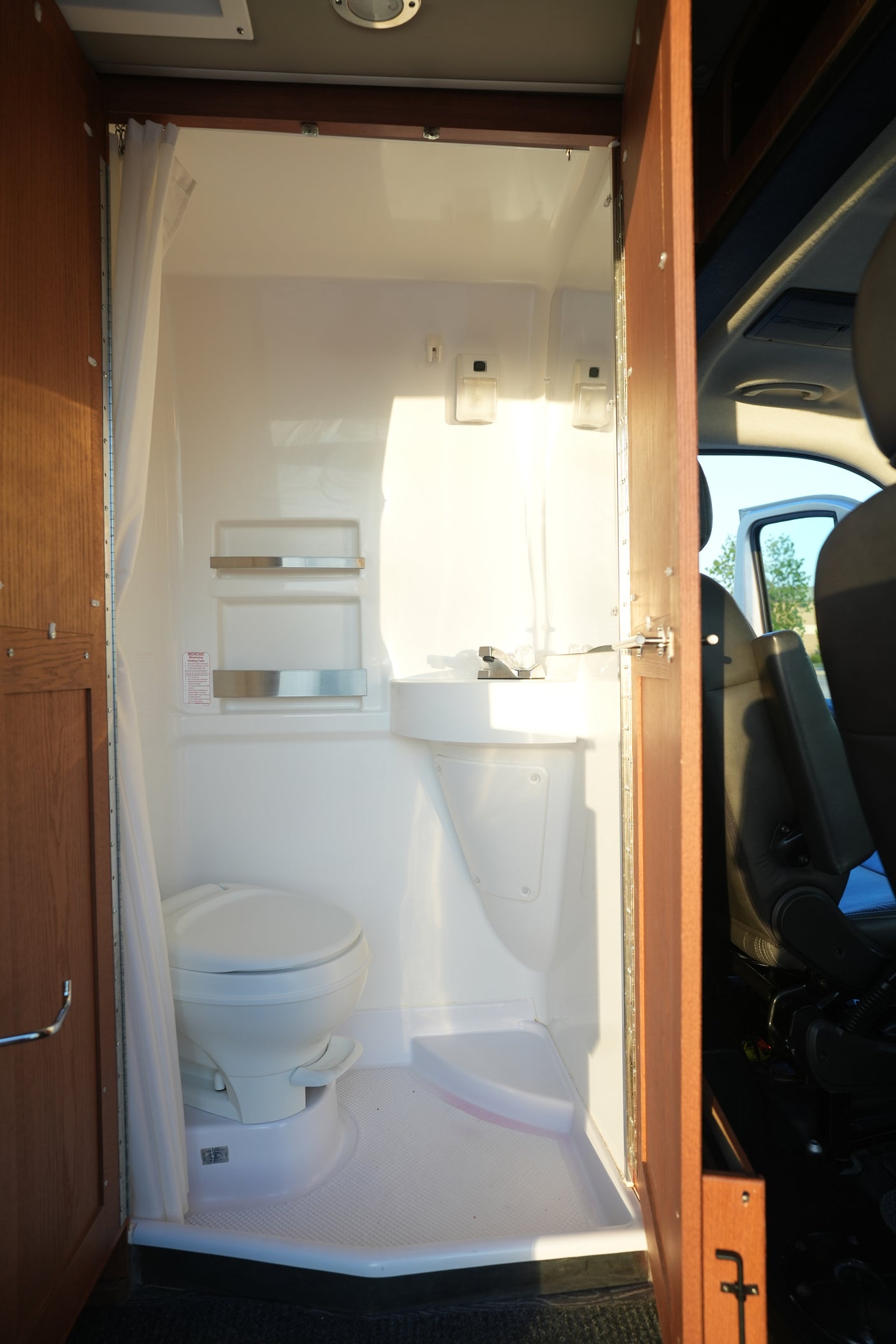 Inside an RV rental: a compact wet bath with toilet, corner sink, and shower curtain. Bright sunlight illuminates the clean, functional space.