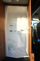Inside an RV rental: a compact wet bath with toilet, corner sink, and shower curtain. Bright sunlight illuminates the clean, functional space.