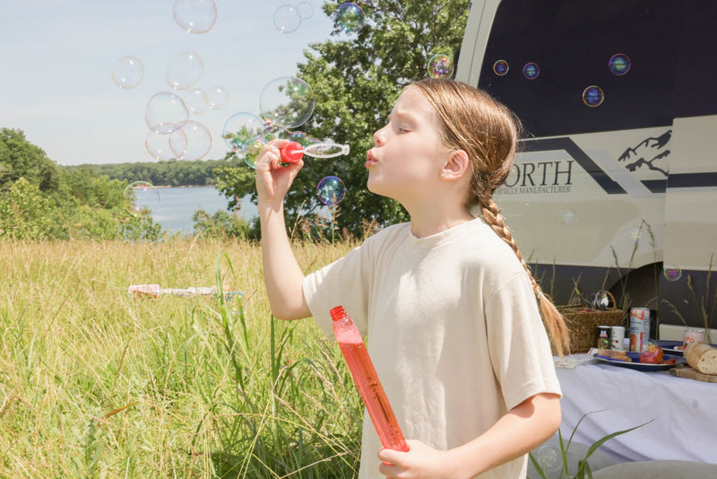 A young girl blows colorful bubbles outdoors by an RV rental, with a lake and trees in the background. — photo 13