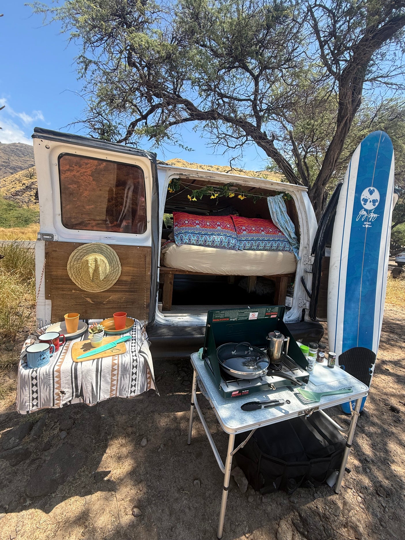 This rv rental campervan, ready for adventure, features an open back with a bed, an outdoor kitchen with stove, and a surfboard.