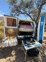 This rv rental campervan, ready for adventure, features an open back with a bed, an outdoor kitchen with stove, and a surfboard.