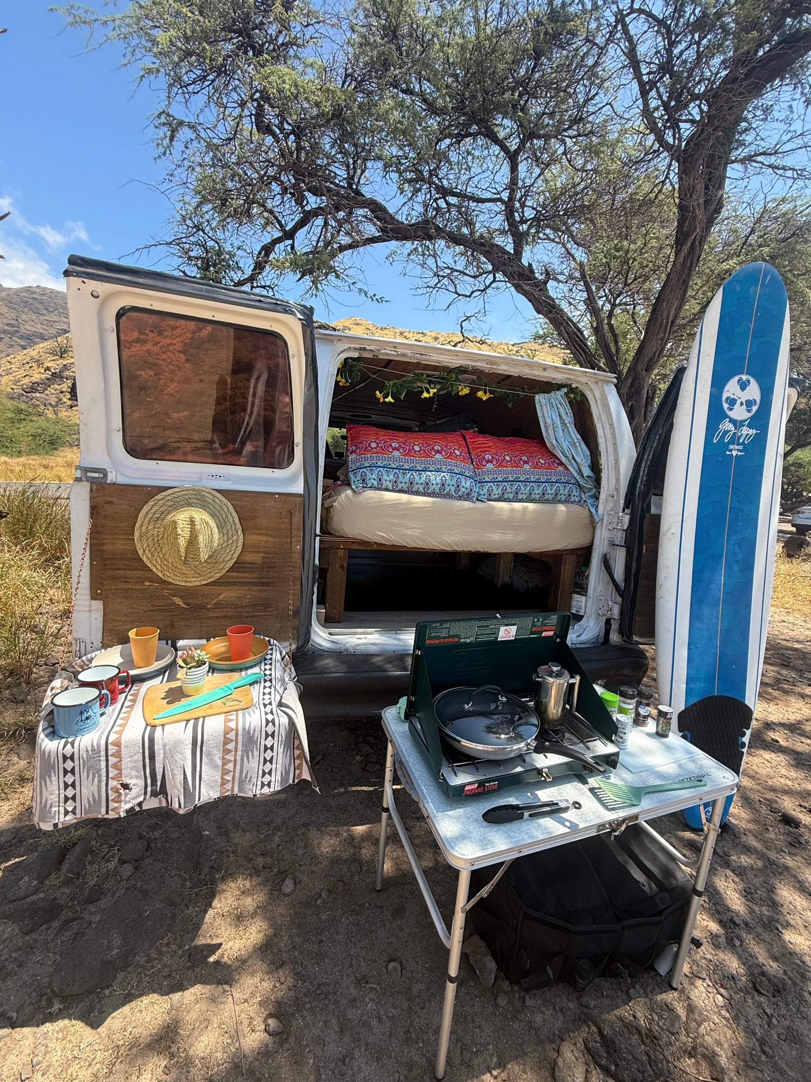 This rv rental campervan, ready for adventure, features an open back with a bed, an outdoor kitchen with stove, and a surfboard.
