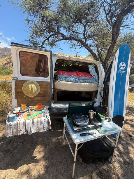 This rv rental campervan, ready for adventure, features an open back with a bed, an outdoor kitchen with stove, and a surfboard.
