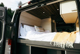Cozy, white-sheeted bed with a yellow throw inside an RV rental camper van, seen from the open rear doors, hinting at comfortable mobile living.