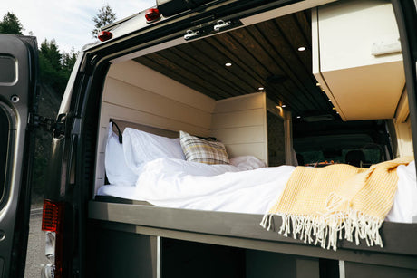 Cozy, white-sheeted bed with a yellow throw inside an RV rental camper van, seen from the open rear doors, hinting at comfortable mobile living.
