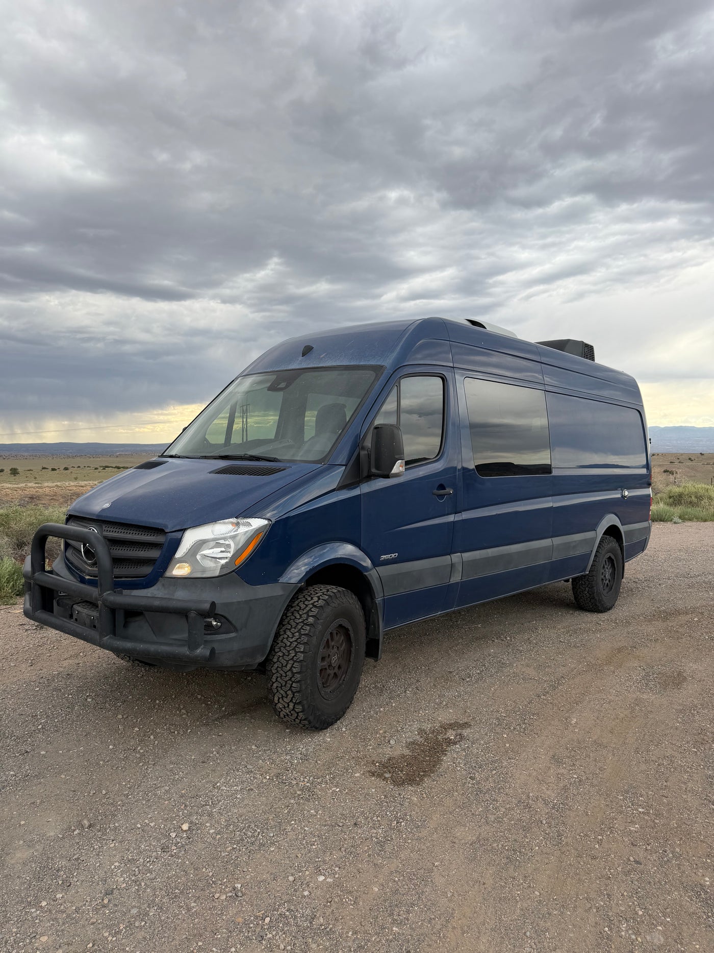 Stylish blue Mercedes Sprinter rv rental with off-road tires, parked on a dusty path under an overcast sky, ready for adventure. — photo 1