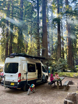 RV rental parked in a majestic Redwood forest campsite. Sunlit rays pierce the canopy as two girls prepare food at a picnic table.