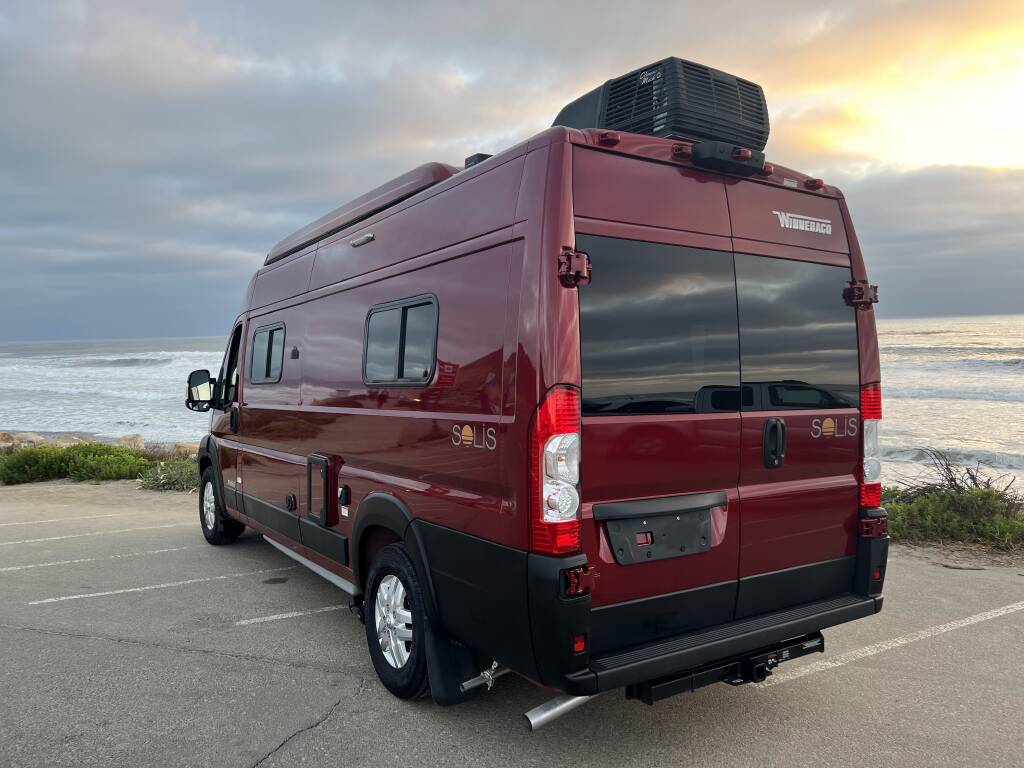 Red Winnebago Solis rv rental camper van by the ocean. Rear view showing side windows, rooftop AC, and marine backdrop.